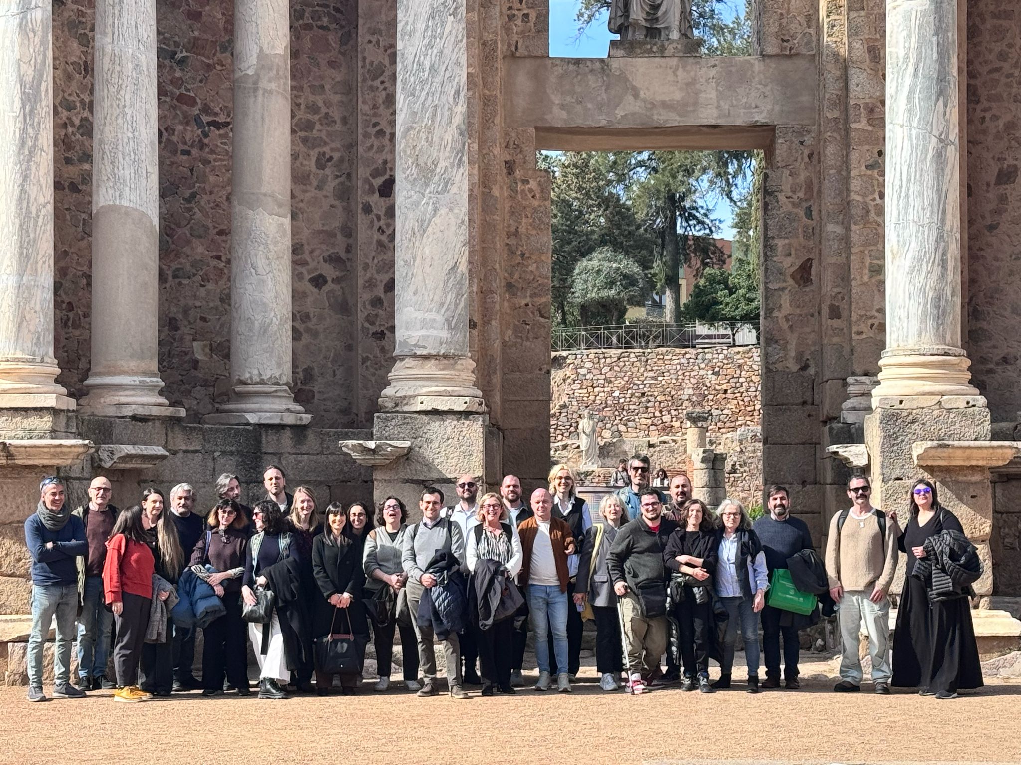 The image shows a large group of people, approximately 30 individuals, posing for a group photograph in front of the ruins of an ancient structure. The architecture appears to be Roman or Greco-Roman in style, featuring large, weathered columns and stonework. The group is diverse in age and attire, suggesting a possible tour group or academic gathering. The setting is outdoors, on a sunny day. The overall impression is one of a cultural excursion or historical study visit.