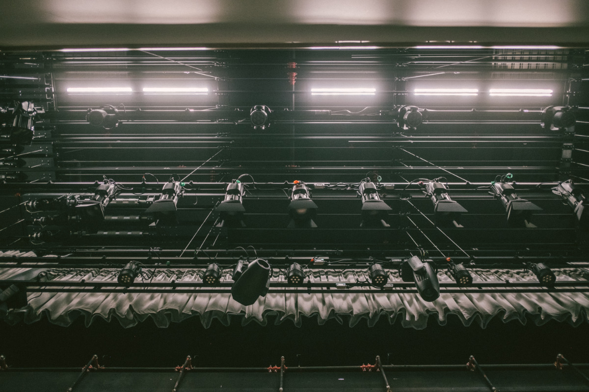 The image shows a low-angle view of the lighting grid above a stage. Numerous spotlights and long fluorescent lights are suspended from a complex network of metal bars and wires. A curtain is visible at the bottom of the frame. The overall atmosphere is dark and industrial, emphasizing the technical aspects of stage production.