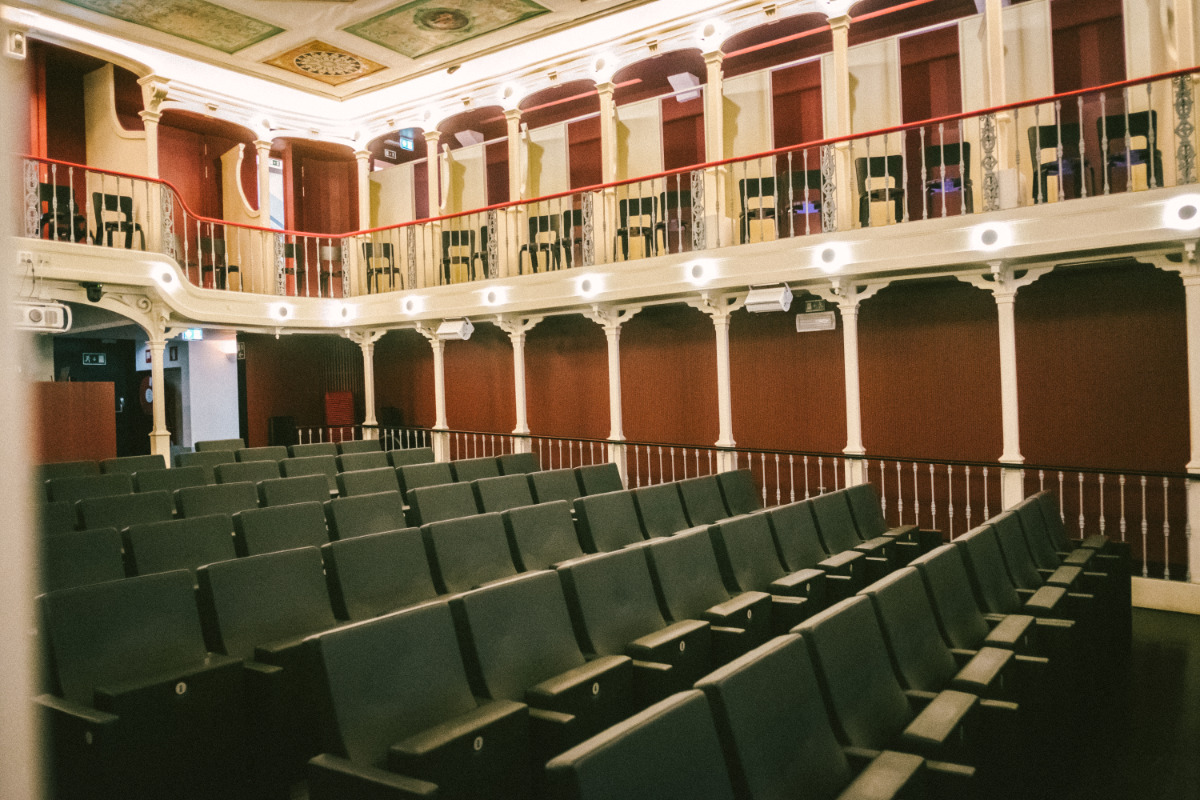 The image shows the interior of a theater or auditorium. The seating area is filled with rows of dark gray upholstered chairs. A balcony with chairs and railings is visible above the main seating area. The architecture is ornate, with painted ceilings and classical columns supporting the balcony. The overall color scheme is muted and dark, with a reddish-brown tone to the walls. The scene is empty, giving the impression of a quiet, awaiting-audience atmosphere inside São Luiz